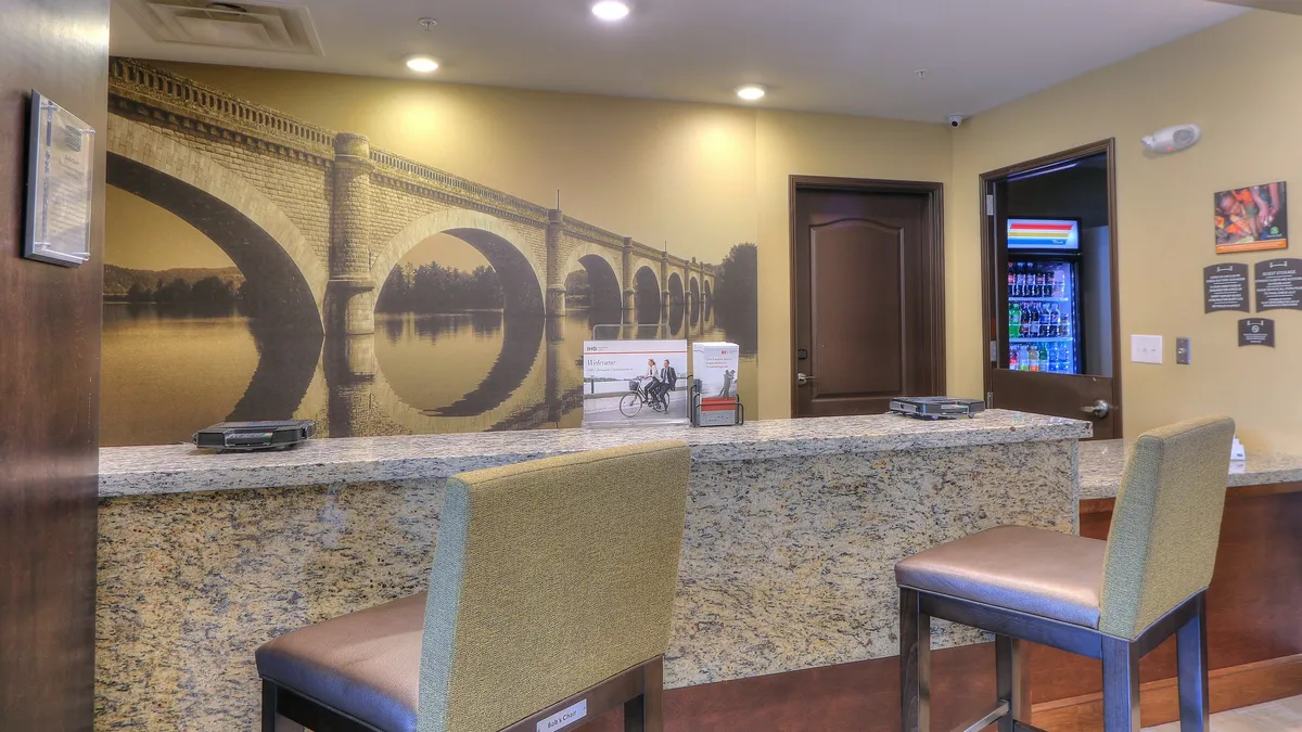 The image captures the lobby area, featuring a warm granite countertop, inviting reception chairs, and a mural of a local bridge that subtly connects the property to its surrounding community. Guests can be seen checking in, interacting with friendly staff, and accessing a vending machine for quick essentials.