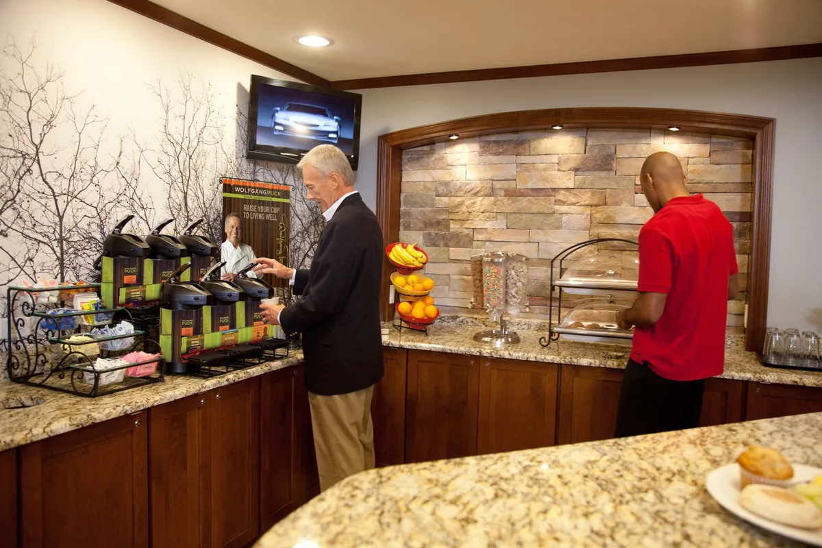 This image depicts a well-lit kitchen area with granite countertops, coffee machines, a stone accent wall, and a vibrant fruit display. The setting feels inviting and practical, with easy access to morning beverages and snacks, complemented by the contemporary decor and functional workspace.