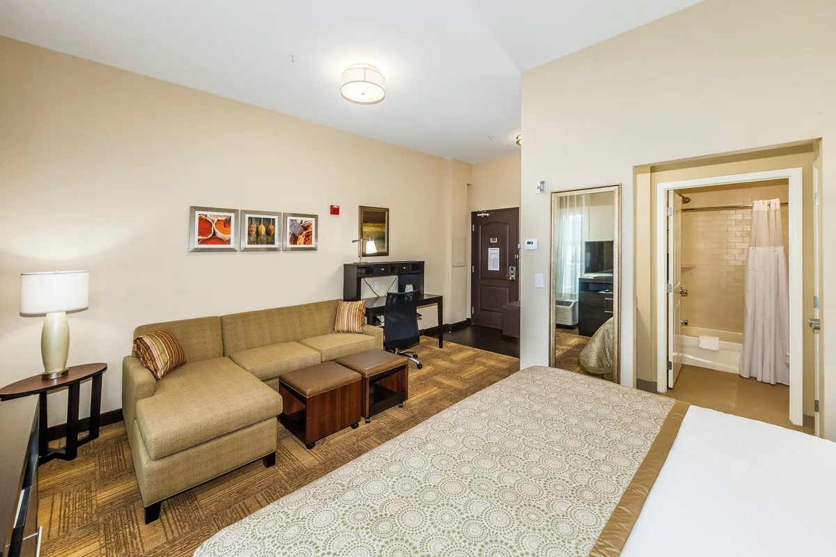 The image showcases a modern studio interior with a comfortable bed adorned in patterned bedding, a cozy sofa seating area perfect for relaxation, tasteful framed artwork on the wall, and a glimpse of the bathroom entrance. The space feels bright, inviting, and thoughtfully designed for both rest and productivity.