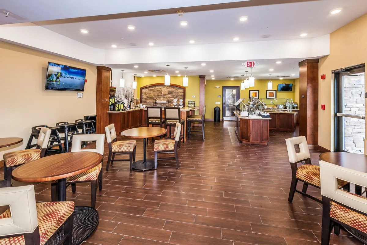 The scene shows a cozy coffee bar adjacent to a seating area, with a decorative stone wall and a television screen. Guests can be seen enjoying a casual dining setup, which doubles as a space for socializing, working, or watching TV. The furniture and space organization suggest flexibility for dining or lounging.
