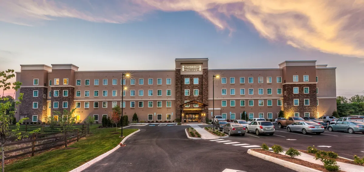The image captures the exterior of the hotel, featuring a welcoming entrance framed by well-maintained landscaping and visible parked cars. The scene hints at social gatherings and the flow of business and leisure guests entering and exiting the property.