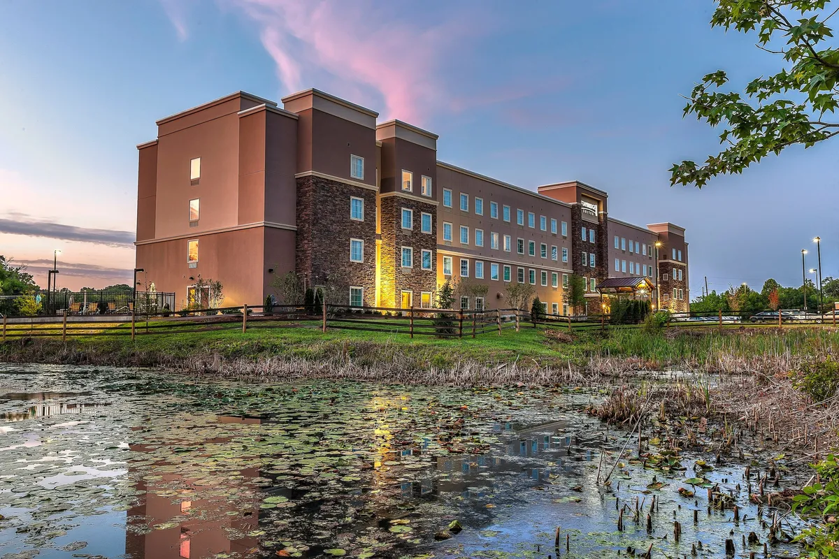 This exterior scene captures the building’s facade reflected in a tranquil pond, surrounded by natural landscaping. The peaceful setting and sunset colors enhance the sense of a relaxing retreat connected to nature and open spaces.